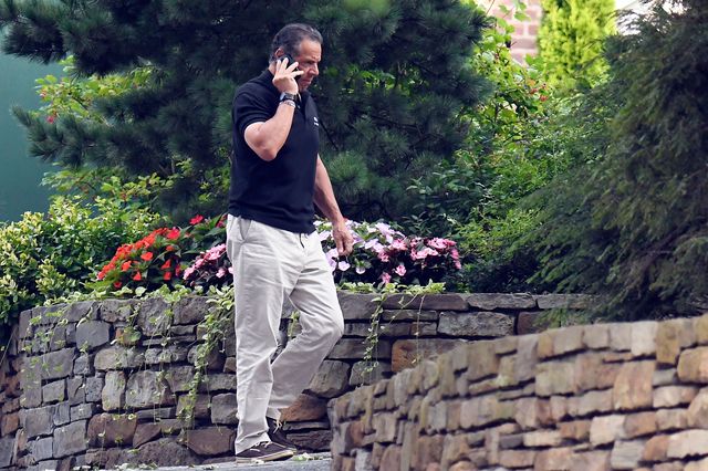 Governor Cuomo, in a navy blue polo shirt and khakis, walking outside by a stone-walled garden